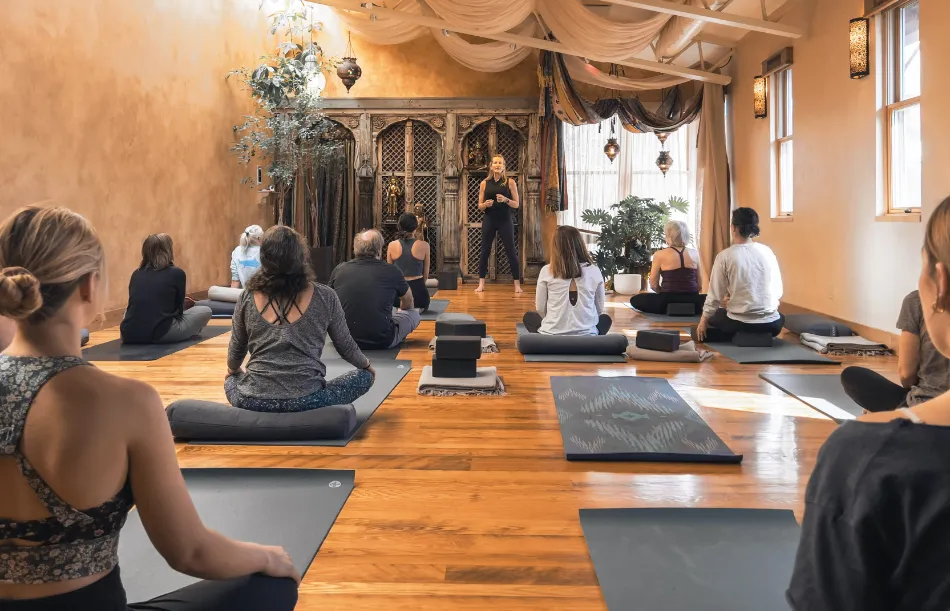 View from the back of the room of a yoga teacher teaching a yoga class with several students seated facing forward and paying attention