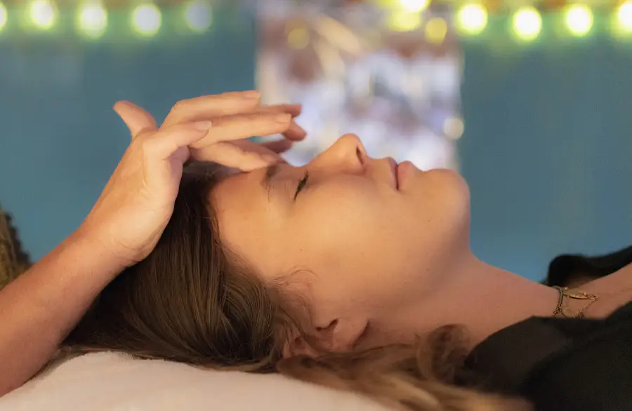 Image of a woman's side profile as she lays down on a massage table while another woman puts her fingers in between her eye brows as she performs Reiki.