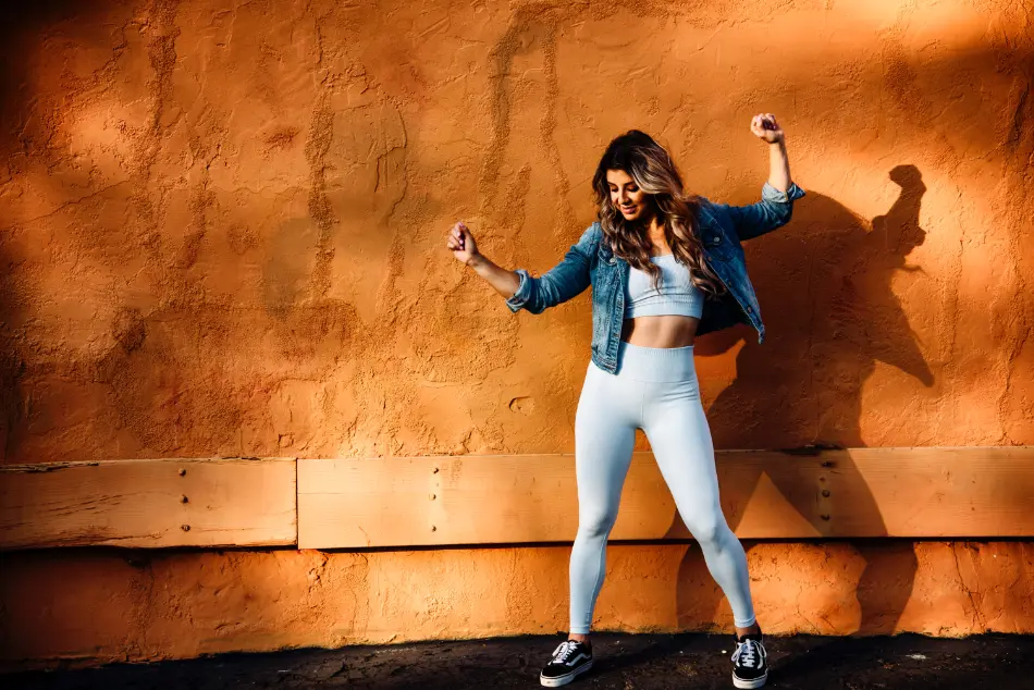 Woman wearing a tank top, jeans jacket, and leggings yoga pants in front of an orange wall