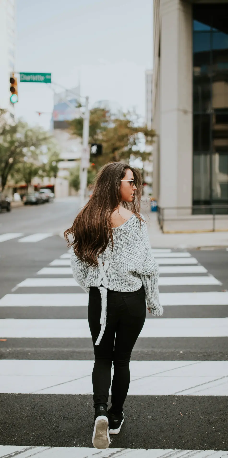 Woman wearing a gray sweater, with black leggings yoga pants, and sneakers, crossing a zebra striped crosswalk and looking over her right shoulder