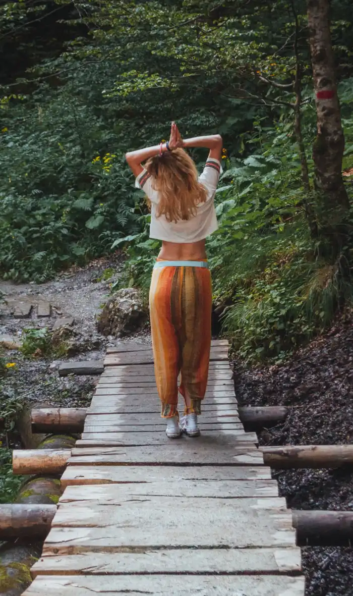 Back view of a woman in flowy yoga pants with her arms raised above her head, palms together, standing on a wooden bridge in what looks to be a lush national park with ferns and green foliage.