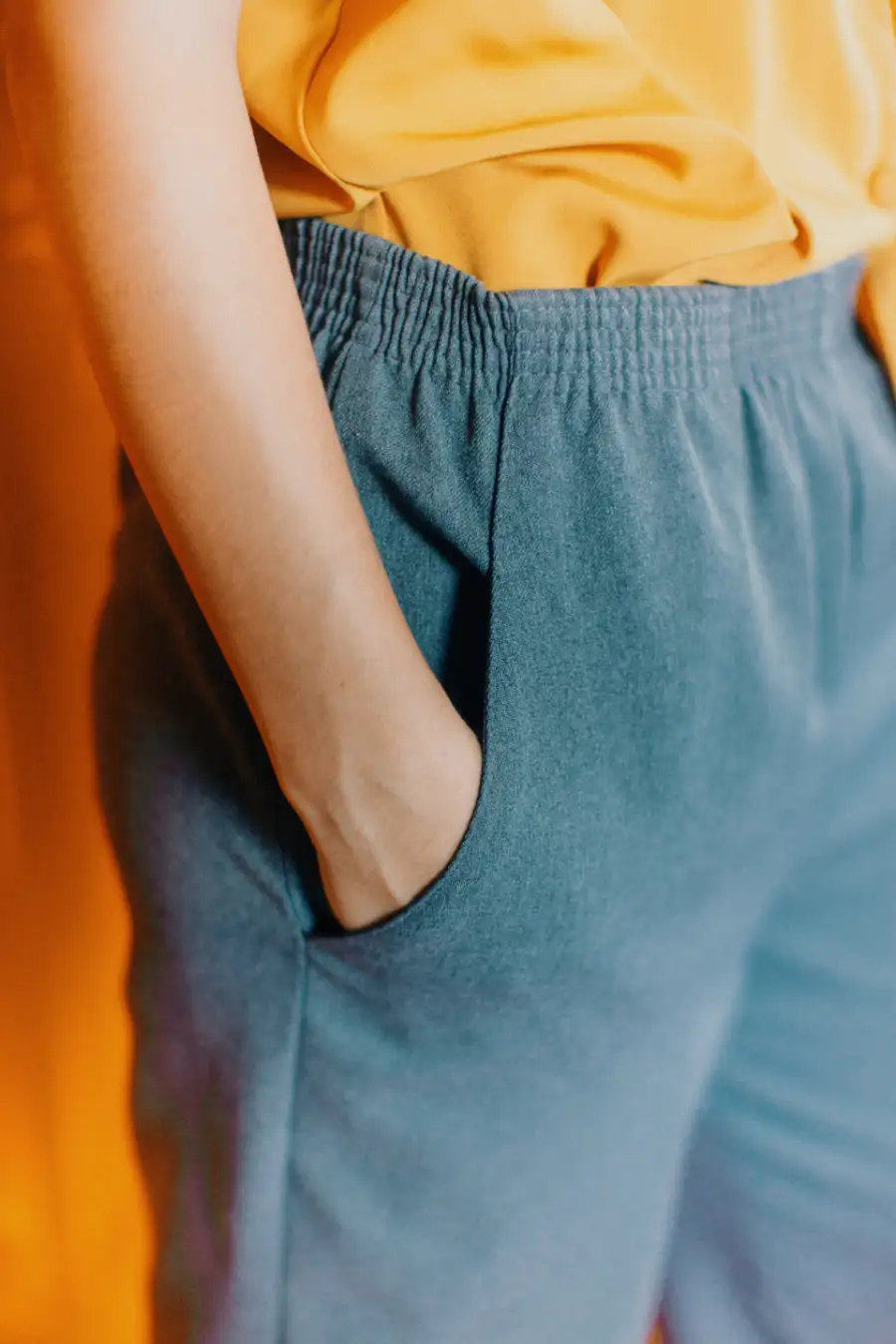 View of the mid section of a person. You can see the bottom of their orange shirt and the tops of their gray, jogger yoga pants. They are placing their hands in their pockets.