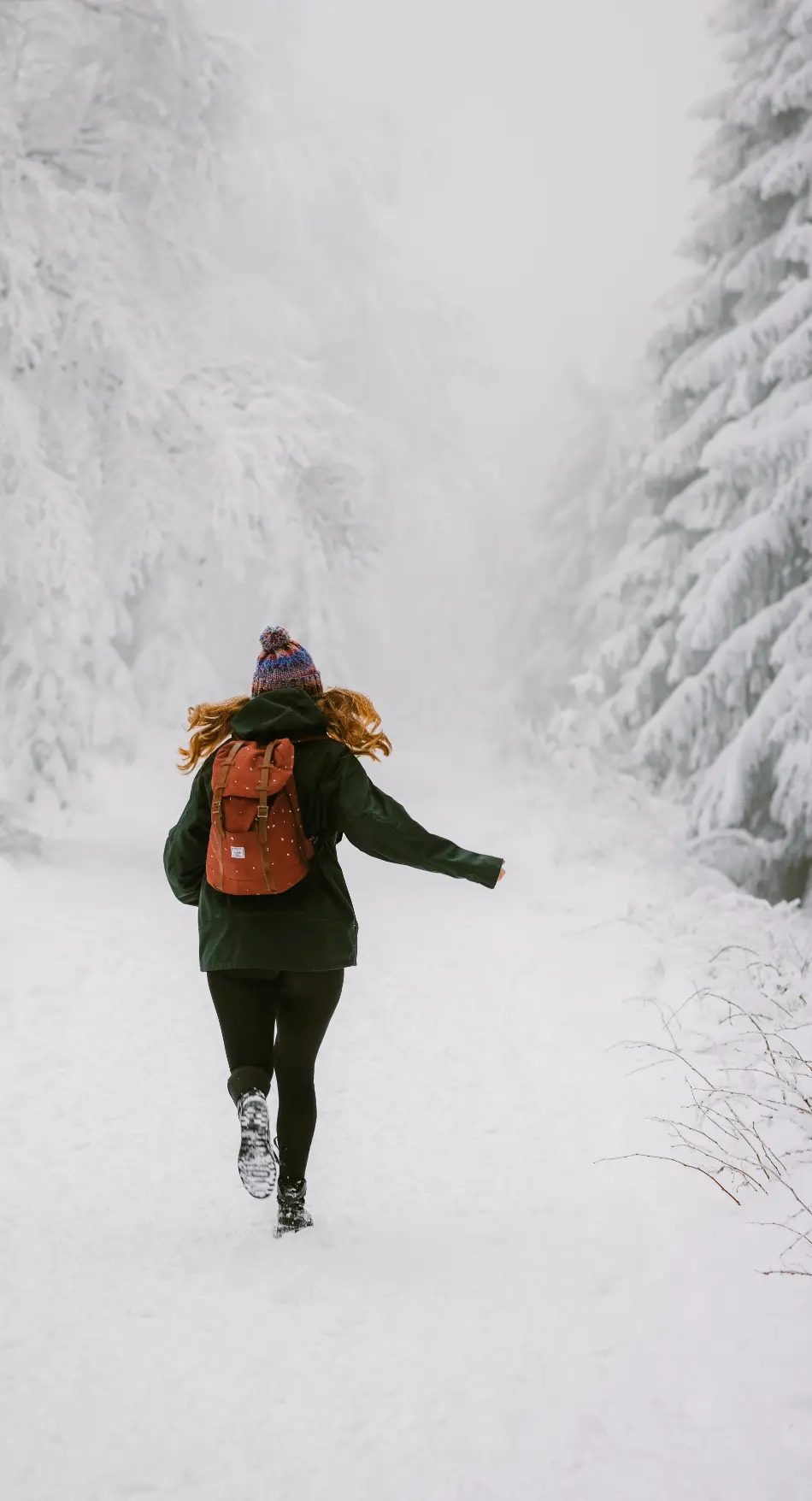 View from behind of a woman running through the snow. She has a jacket, backpack, beanie, black leggings and hiking shoes.