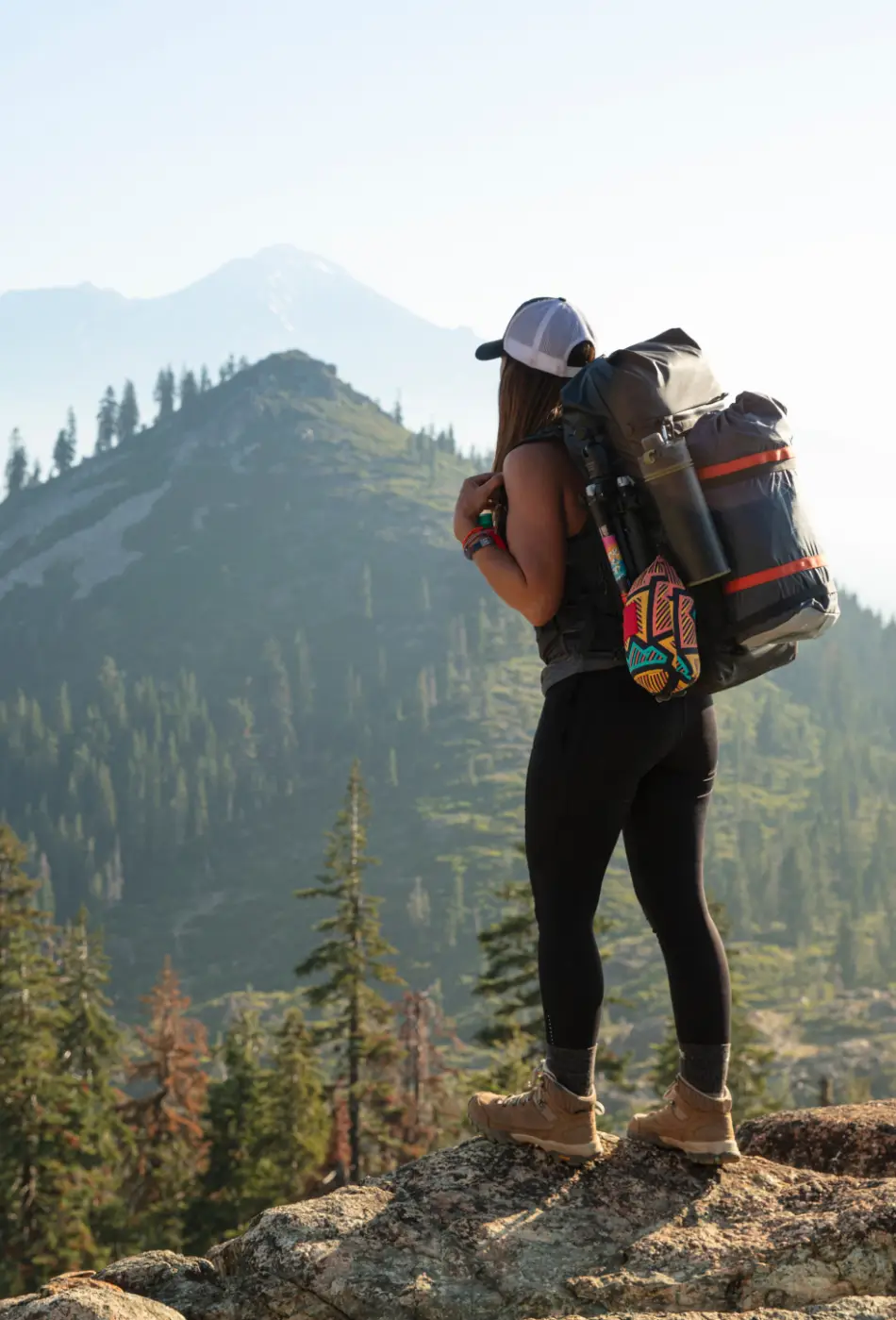 View of a woman as she looks the horizon. She has on her back a very large back pack with various day hike items. She is wearing a trucker baseball hat, black leggings and hiking boots.