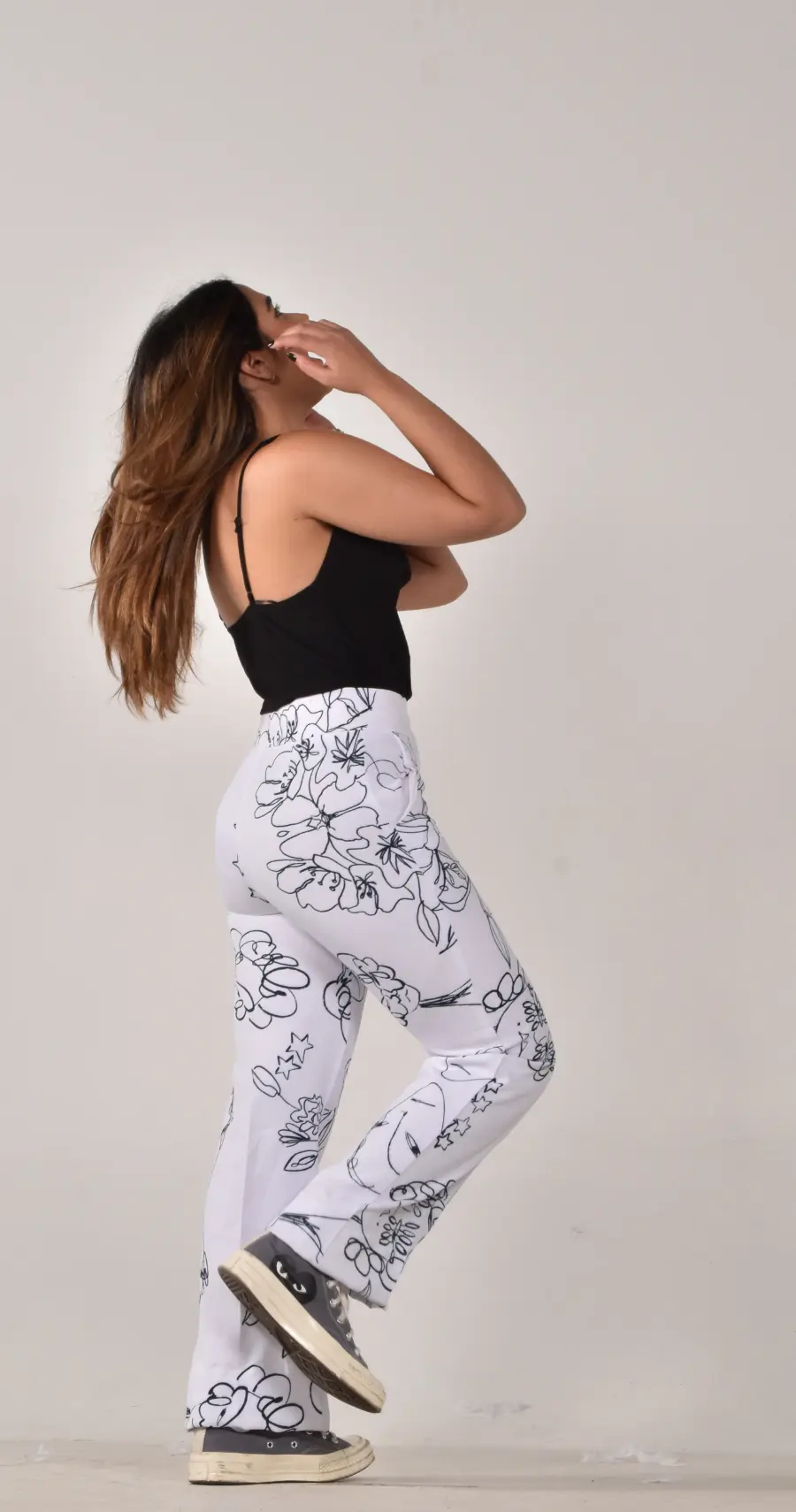 Side view of a woman wearing a black tank top and white yoga pants with simple black outlined floral print. She is brushing her hair back and looking up towards the ceiling.