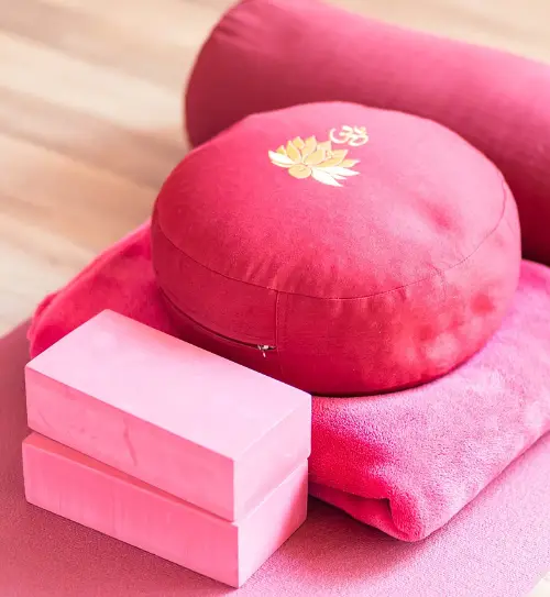 Image of yoga accessories: A towel, yoga blocks, a round seating pillow and a head rest tubular pillow. They are all colored variations of pink. The seating pillow has a gold Om (Aum) symbol and lotus flower symbol on it.