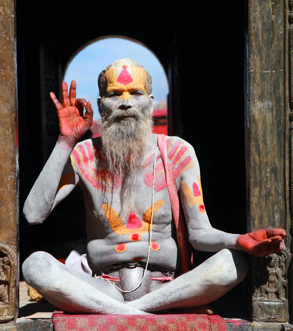 Indian yogi with long beard inside a temple door makes a mudra with his hands. His body is painted with red and yellow on his dark skin.